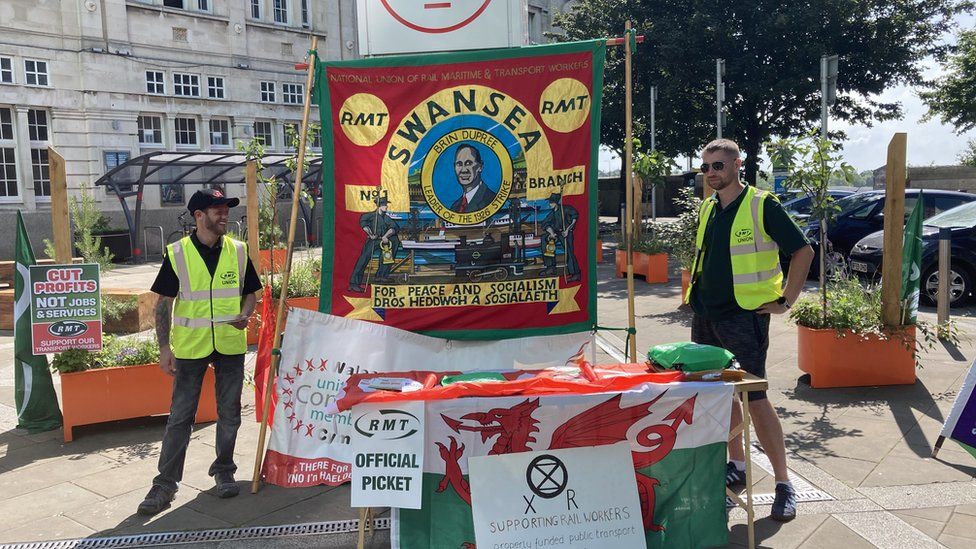 Two members of the RMT union in hi-vis vests stand beside posters at the picket line