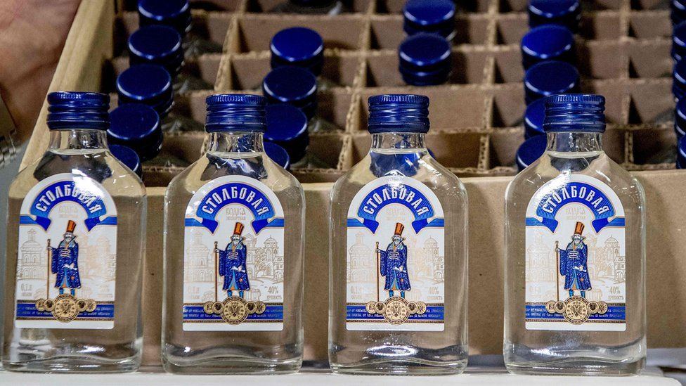 Picture shows vodka bottles with blue caps and writing in Russian, sitting in their cardboard boxes