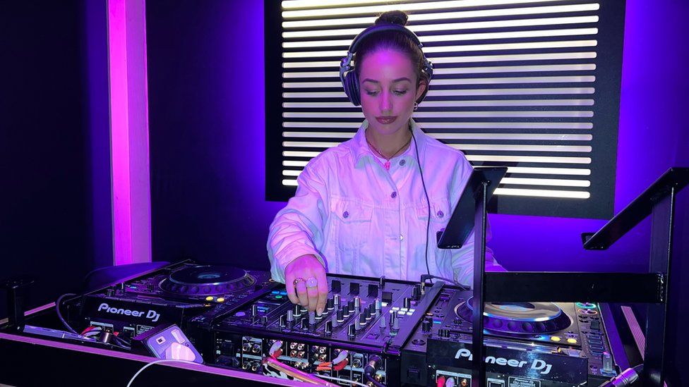 Hannah Rose is in a studio behind a Pioneer mixing desk. She has two turntables in front of her, and is adjusting a dial on a mixer with rows of different adjustment knobs. She's looking down at the equipment. Her hair's tied up in a bun, she has over-ear headphones one, but the left cup is pulled behind her ear and not actually covering it. She's wearing a white denim-style jacket, but it's reflecting the neon pink and blue lights that illuminate the studio.
