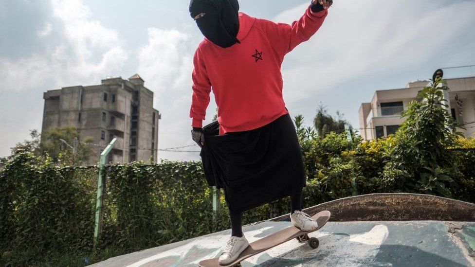 A girl on a skateboard. She is wearing a niqab, a long black dress and a red hoodie.