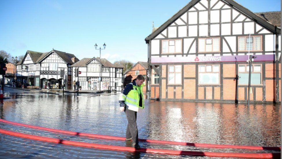 A member of the Environment Agency crosses a flooded road walking on pipes