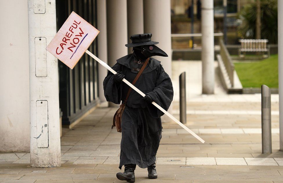 A man in a 17th Century plague doctor"s costume carries a sign in central Leeds on March 21, 2020, a day after the British government said it would help cover the wages of people hit by the coronavirus outbreak as it tightened restrictions to curb the spread of the disease.