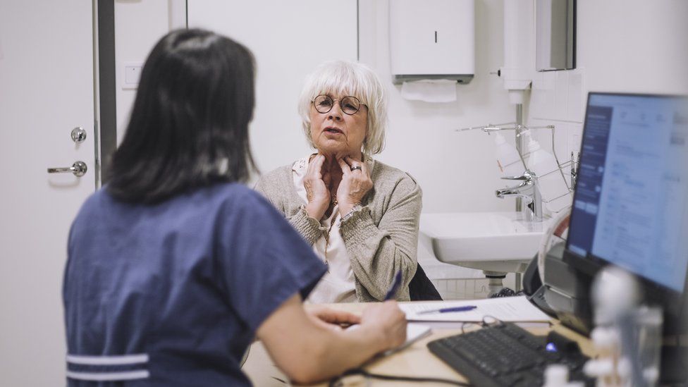 Older female patient sits opposite female doctor, describing her symptoms of neck pain