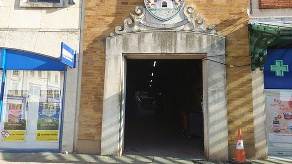 Tunnel-like entrance to walkway between shops, with crest above
