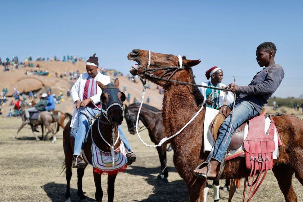 Men and a boy on brown horses. In the background there are hills and a crowed of people scattered around.