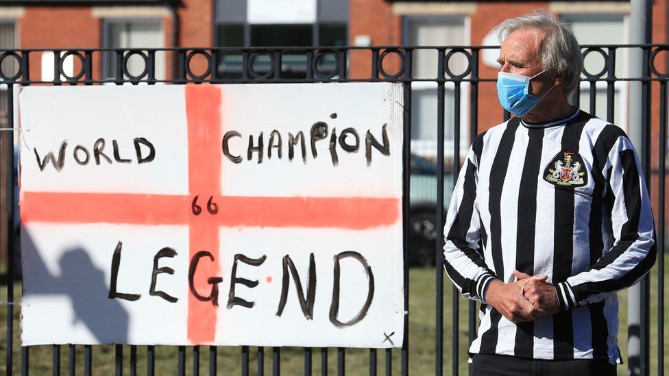 A man in a Newcastle United shirt stands next to a tribute flag celebrating England's 1966 World Cup win