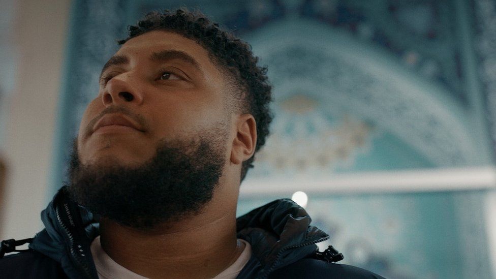 Big Zuu from from his his documentary Big Zuu Goes to Mecca. Big Zuu is a 28-year-old black man with short afro hair, brown eyes and a dark beard. He wears a black jacket over a white T-shirt and is pictured inside a mosque decorated in a rich blue