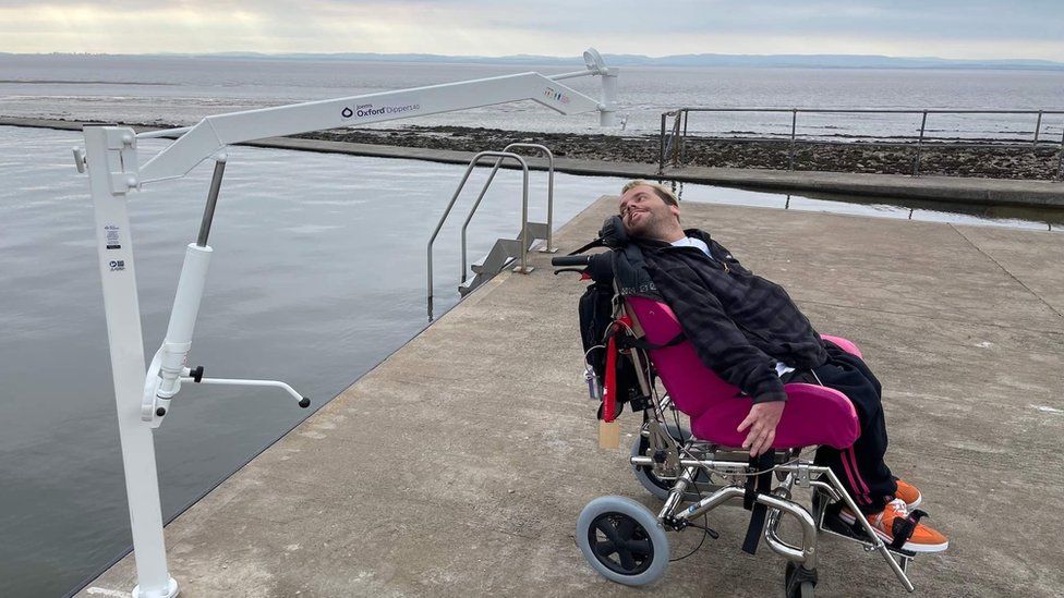 Man in a wheelchair by an electronic hoist at a lake