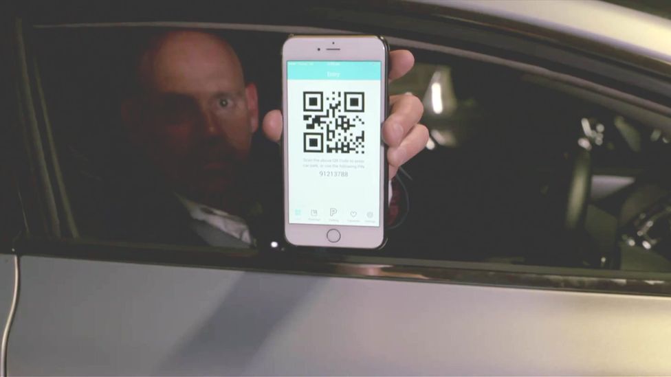 Bald man holding a phone with a code on its screen out of of his car window