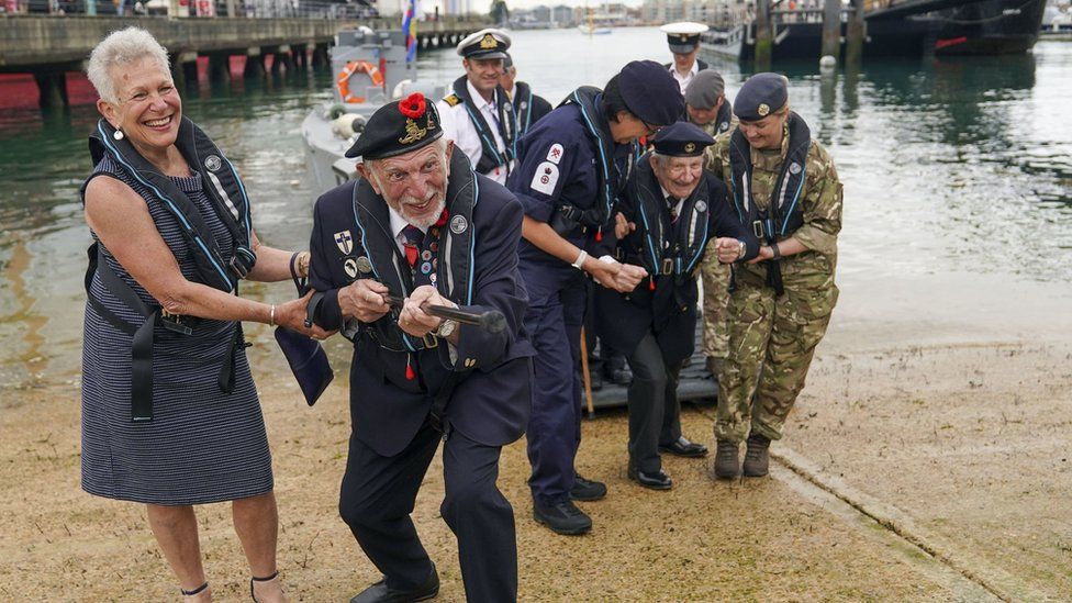Joe Cattini raising his walking stick like a machine gun as he and other veterans are welcomed to the Portsmouth Historic Dockyard to commemorate the 77th anniversary of the Normandy Landings in 2021