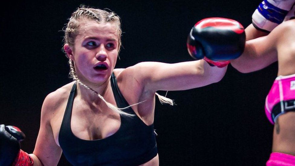 Daniella Hemsley, a blonde white woman with her long hair in braids wears a black sports bra as she aims a punch at opponent Aleksandra Daniel at their fight in Dublin on Saturday 15 July. Aleksandra Daniel wears a pink sports bra with matching shorts and stands with her back to the camera, covering her face with blue and white boxing gloves to defend against Daniella's jab