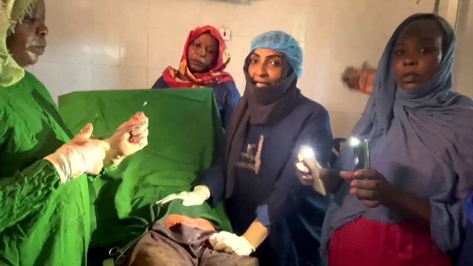 Доктор Ховайда Ахмед аль-Хассан (второй справа) разговаривает с камерой во время кесарева сечения, в то время как женщина освещает пациента светом мобильных телефонов