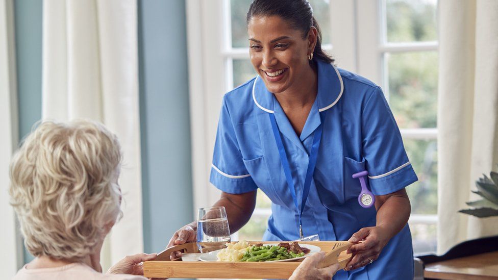 Female Care Worker In Uniform Bringing Meal On Tray To Senior Woman Sitting In Lounge At Home - stock photo