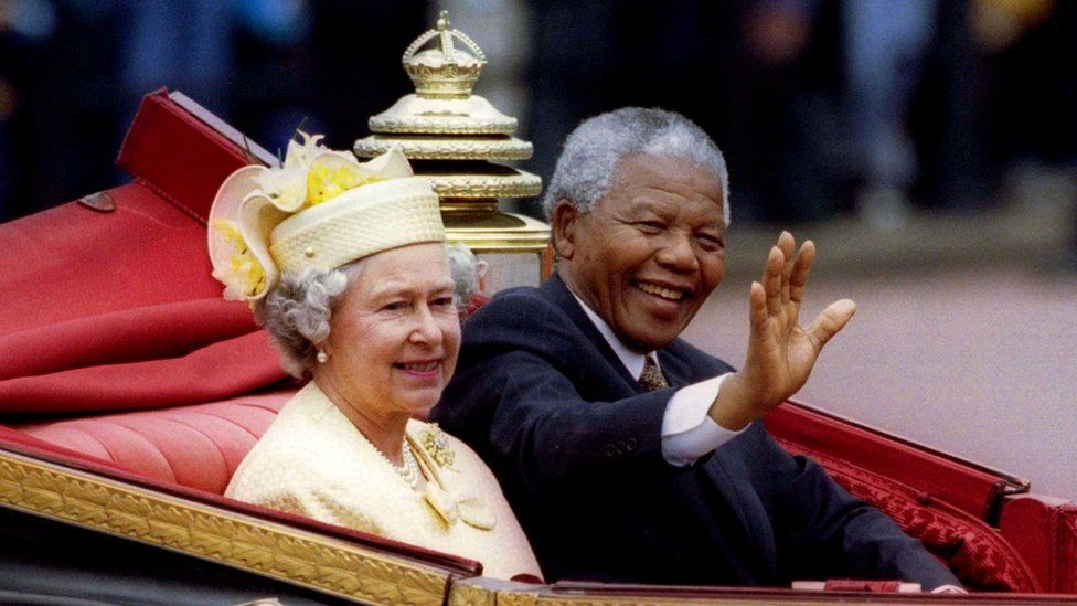Nelson Mandela waves to a crowd while riding in a royal carriage alongside Queen Elizabeth II