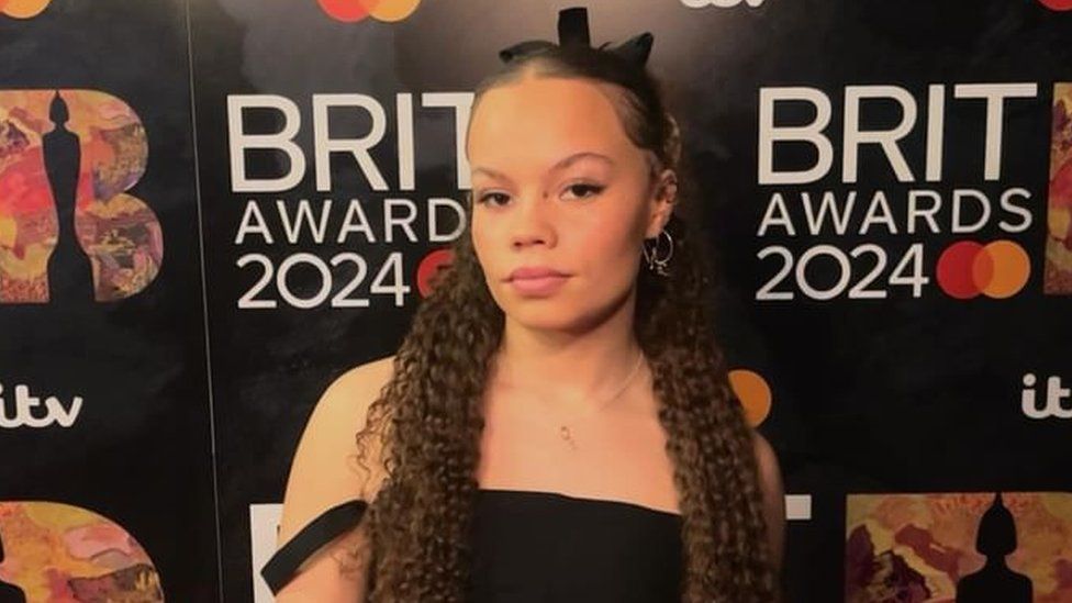 Izzy Withers on the Brits red carpet. Izzy is a 20-year-old mixed race woman with long brown hair. She wears a black ribbon in her hair and a black off-the-shoulder dress.