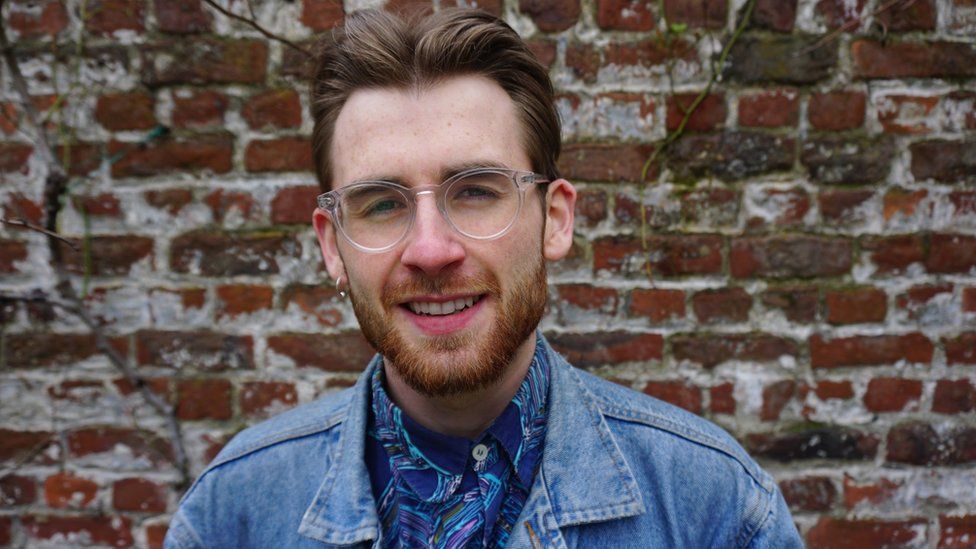 Sean Currie. Sean is a 26-year-old white man with short brown hair and a cropped ginger beard. He wears clear round-framed glasses and has a gold hoop in his right ear. He wears a denim jacket over a patterned blue shirt and is photographed in front of a brick wall.
