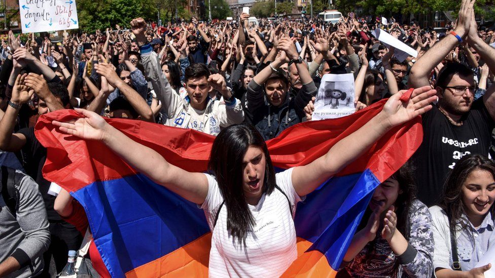 Protesters in Armenia raise their hands, wave flags and hold signs.