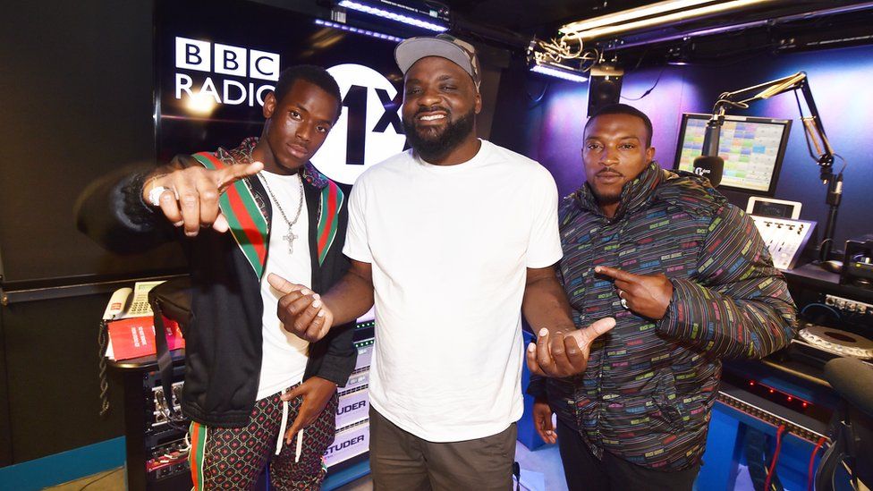 DJ Ace posing in a white T-shirt and cap alongside Top Boy actors Michael Ward, in a black, green, red and white bomber jacket and white shirt and Ashley Walters in a multi-coloured puffer jacket, in the Radio 1Xtra studio.