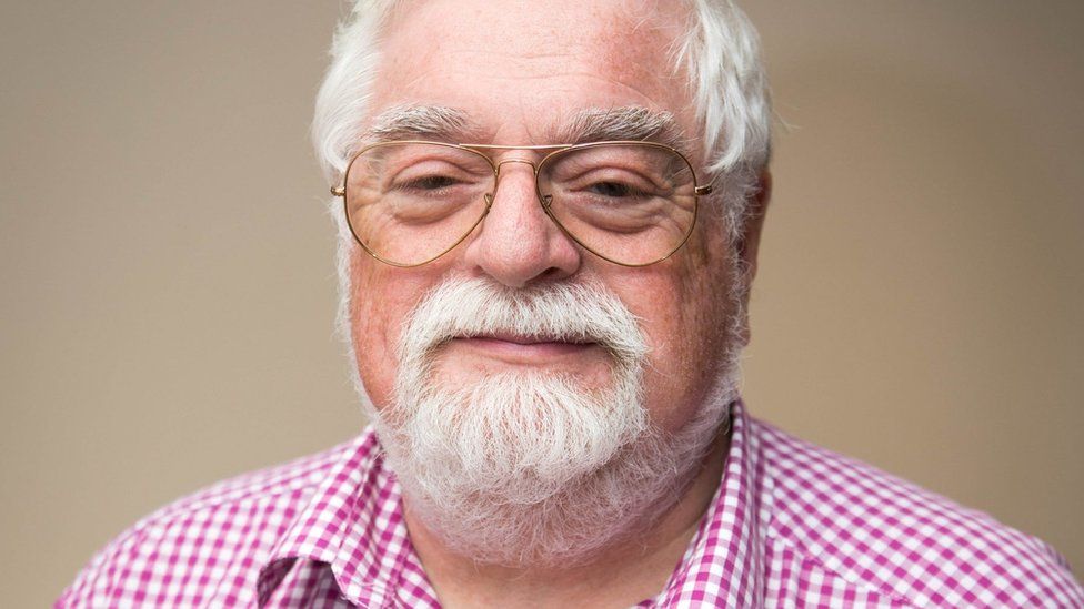 a white man with grey hair and beard and glasses and a pink checked shirt