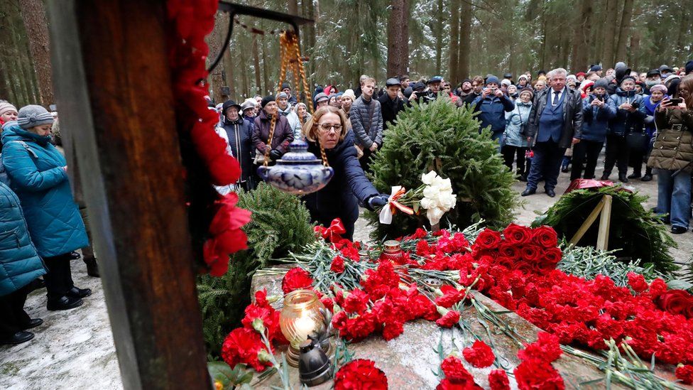 People stand around a mound of wreaths and flowers in Levashovo, with one woman placing a bouquet of white roses on top