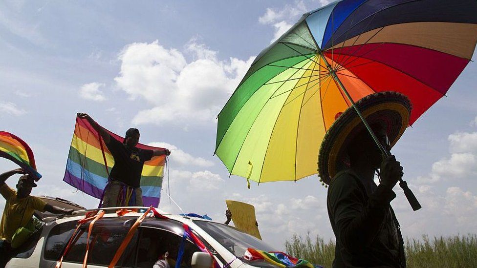 Человек держит зонтик в цветах радужного флага, а другие размахивают флагами во время первого митинга гей-парада после отмены жесткого антигомосексуального закона, против которого власти обжаловали, в Энтеббе, август 2014 года.