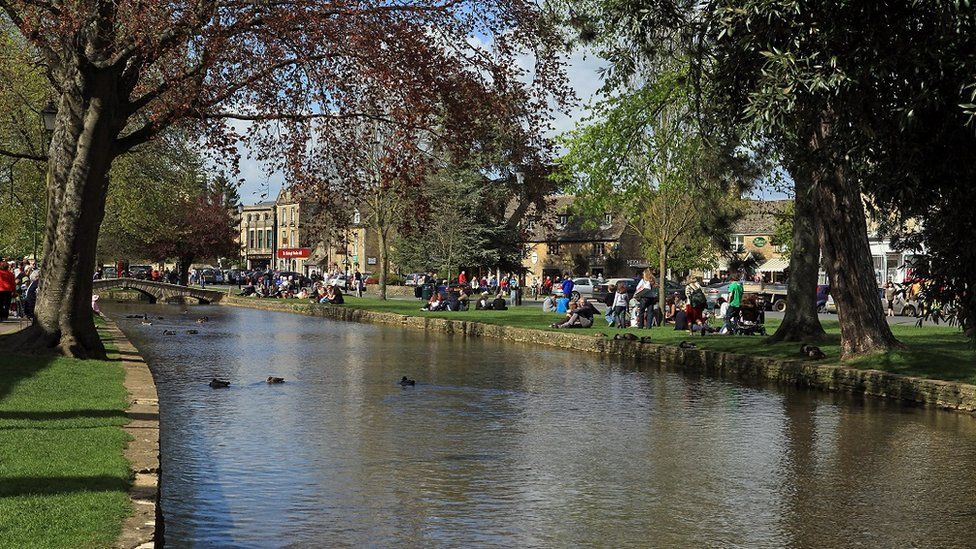 People sitting in the sunshine along the waterside in Bourton-on-the-Water