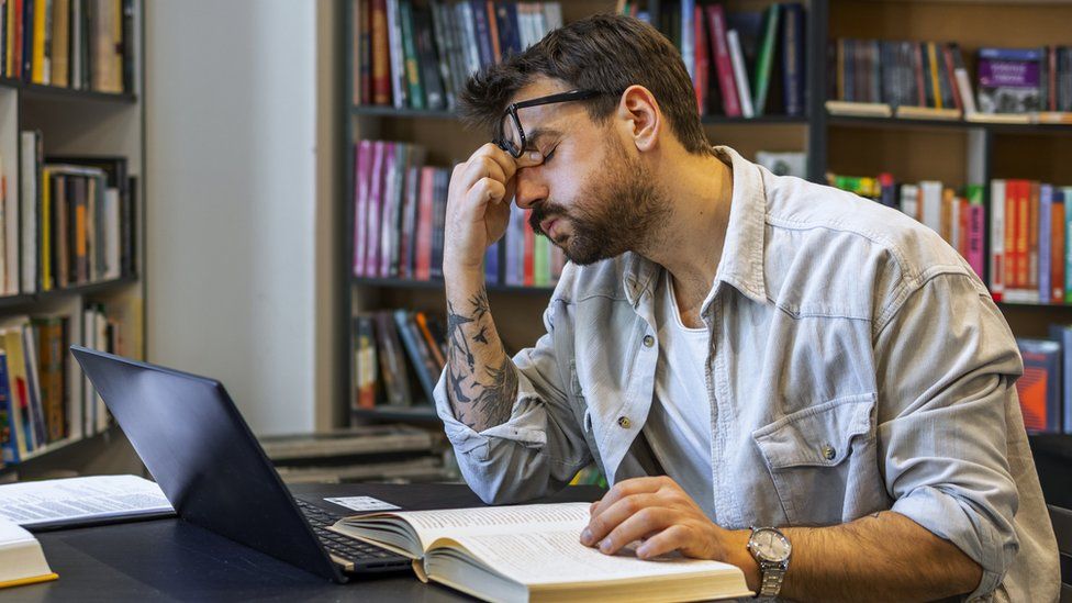 Студент мужского пола выглядит утомленным за столом в библиотеке