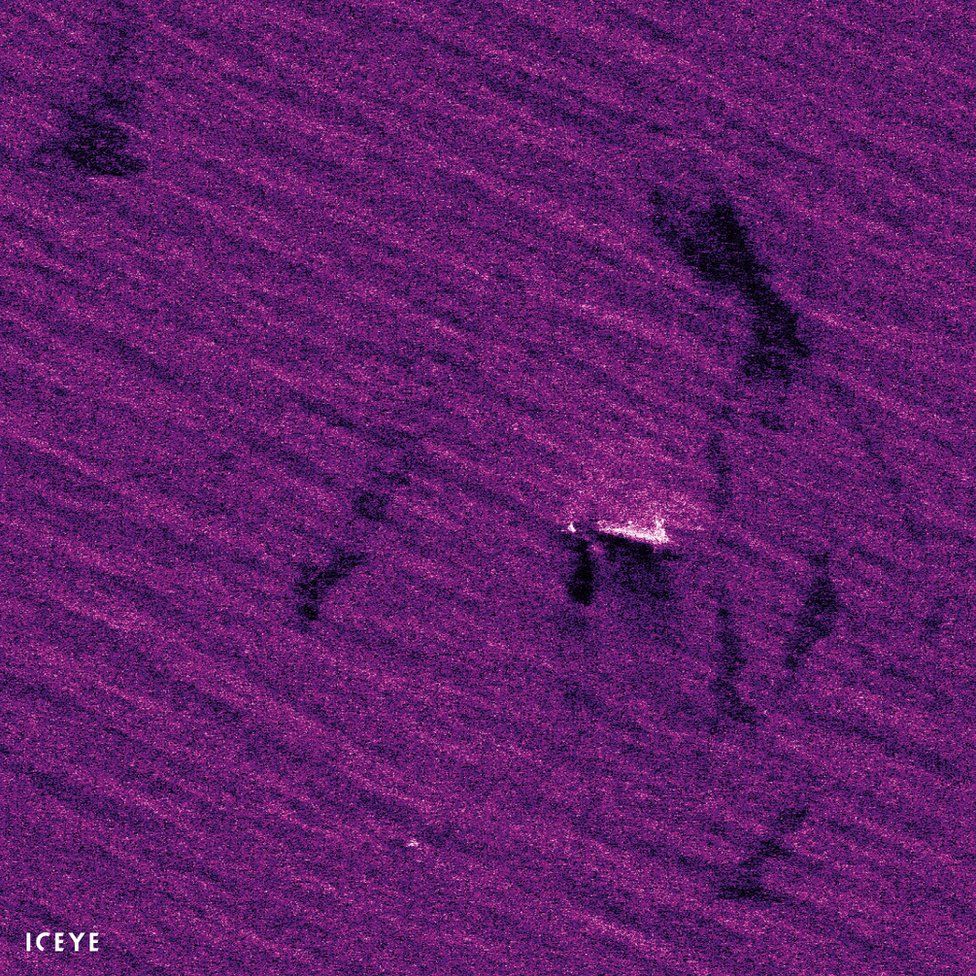 A top-down satellite shot shows a ship upon the ocean, but the colours are unreal and vibrant, with the sea a vivid purple rather than its usual blue - and in this heightened colour state, dark patches can be seen floating on the ocean surface