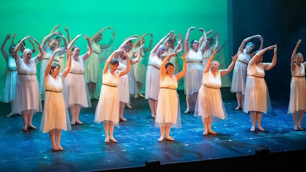 Three lines of women in white dresses hold their hands above their heads while swaying on stage