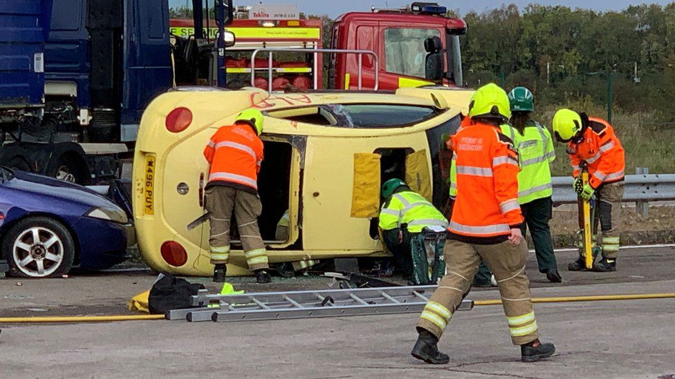 A yellow car on its side in front of a fire truck with five men attending it