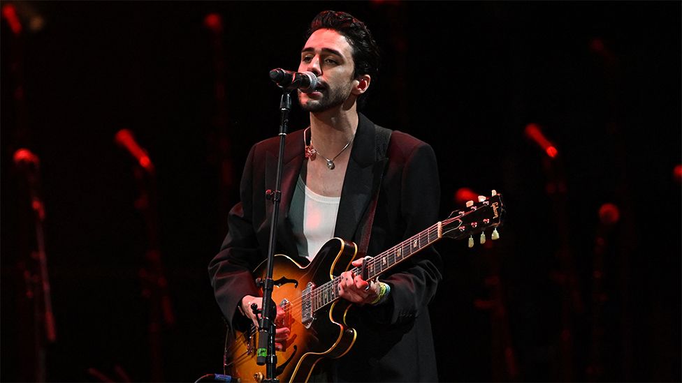 Stephen Sanchez performs at Glastonbury. He's wearing a black blazer with a white vest underneath and a necklace, singing into the microphone. He is holding a guitar which is coloured orange and black. The background has red coloured staging.