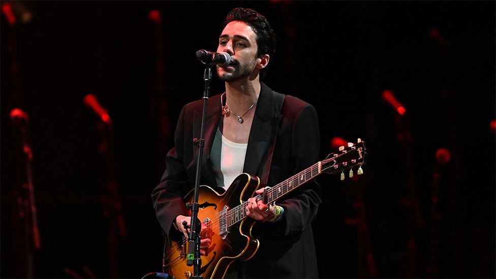 Stephen Sanchez actúa en Glastonbury. Lleva una americana negra con un chaleco blanco debajo y un collar, cantando al micrófono. Sostiene una guitarra de color naranja y negro. El fondo tiene una puesta en escena de color rojo.