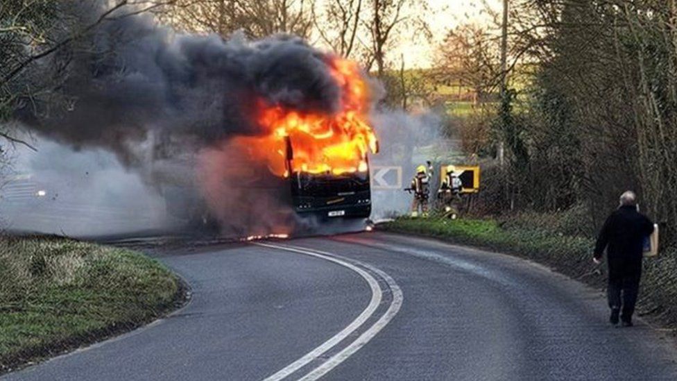 A bus surrounded by flames, with firefighters at the scene