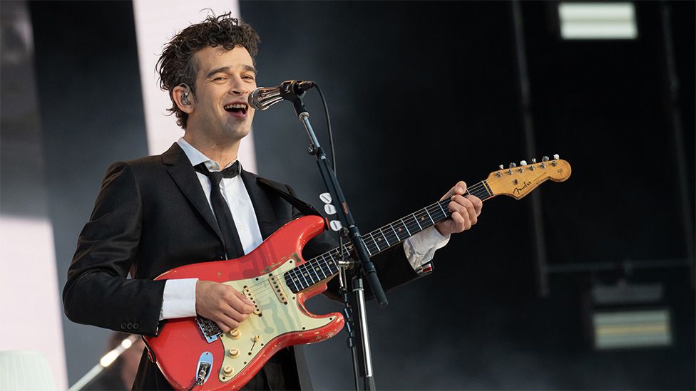 Matty Healy on stage at Radio 1's Big Weekend in Dundee. He's wearing a black jacket and black tie with a white shirt and playing a red guitar.