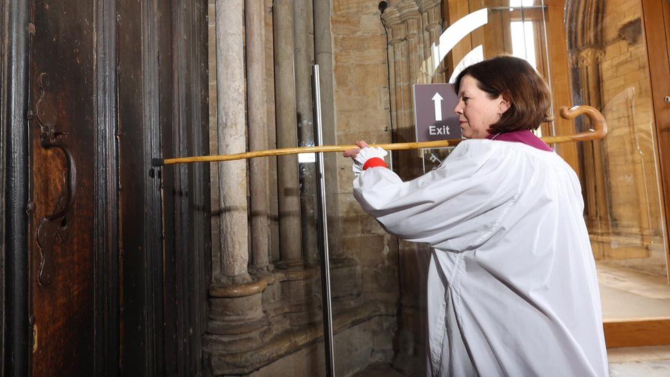 Bishop Debbie knocking on the door of a cathedral with a staff