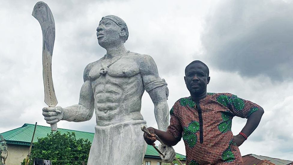 Monday Aigbe standing alongside a statue of his great-grandfather