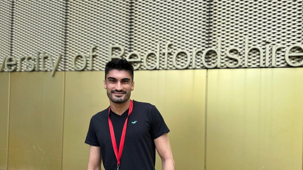 Student Rob Clark wearing a black T-shirt and smiling in front of a gold University of Bedfordshire sign