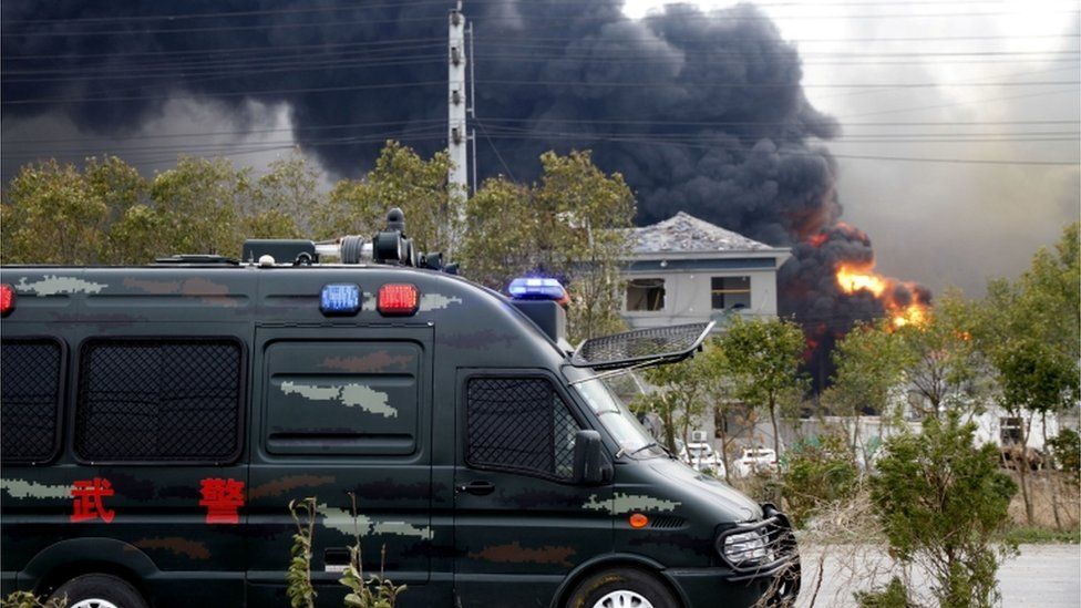 A police van in front of a house with blown out windows, the factory on fire in the background, in Yancheng, China