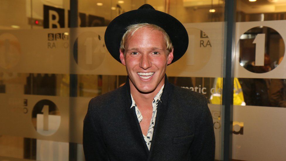 Jamie Laing pictured at BBC Radio 1 offices in 2022. Jamie is a white man with blonde hair in his 30s. He wears a wide-rimmed black hat and a dark coat over a patterned white shirt which is buttoned low. He smiles at the camera and is pictured with BBC Radio 1 branding on windows behind him.