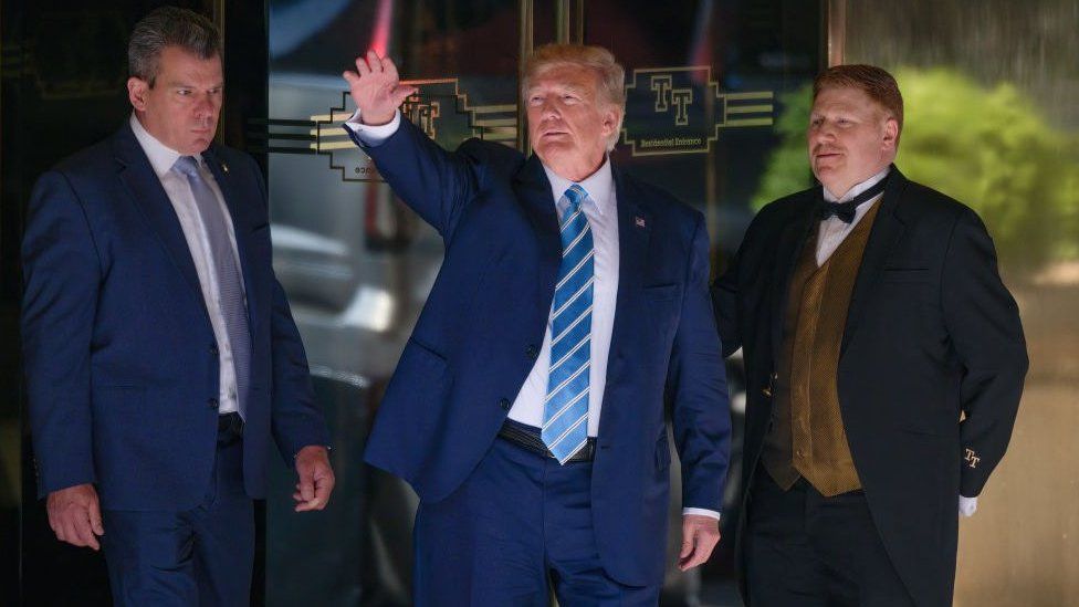 Трамп в синем костюме с синим галстуком перед стеклянными дверями, между мужчиной в синем костюме и швейцаром в черном костюме с золотым жилетом.