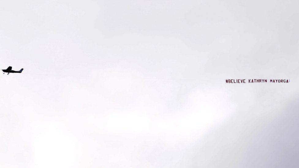 A banner reading "#Believe Kathryn Mayorga" flies over the stadium