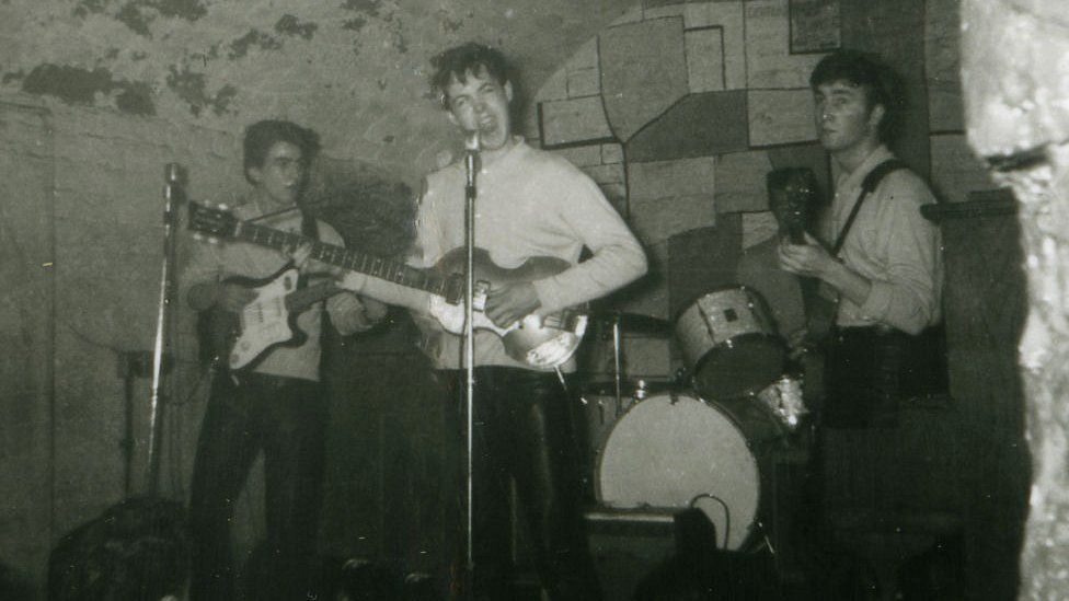 John Lennon and Sir Paul McCartney at the microphone, with George Harrison on guitar and a partially obscured Pete Best, the group's original drummer