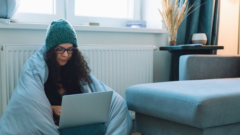 Women sits by radiator working from home wrapped in a duvet