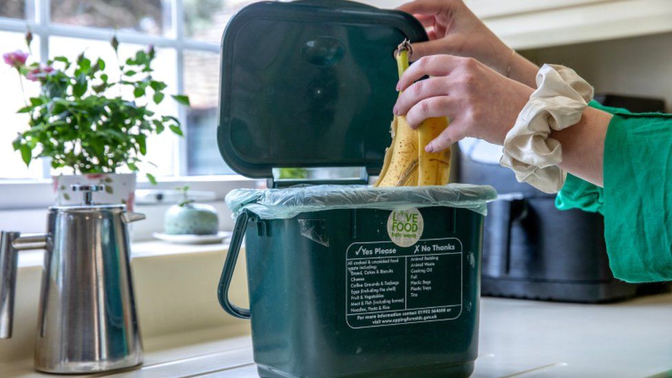 A woman putting a banana skin in a green plastic food caddy with a liner in a kitchen