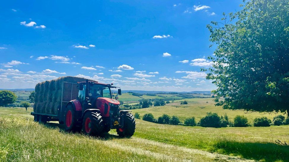 A tractor carrying hay bales driving across a green field with a bright blue sky overhead.