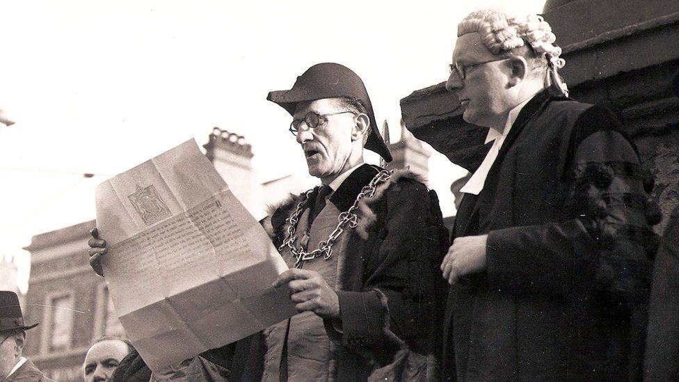 Two men in formal gowns and headwear read a large document in 1952