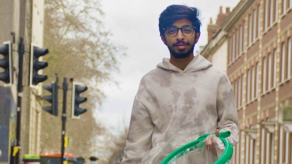 Vivek is stood up holding a litter picker and bag in a built-up area of Bristol