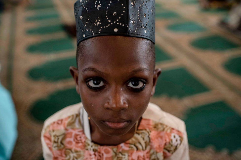 A young boy looks on during a special Jumu'ah prayer service at the Central Mosque in Lagos, Nigeria. He is wearing a hat and his eyes are lined in kohl - Friday 15 February 2019