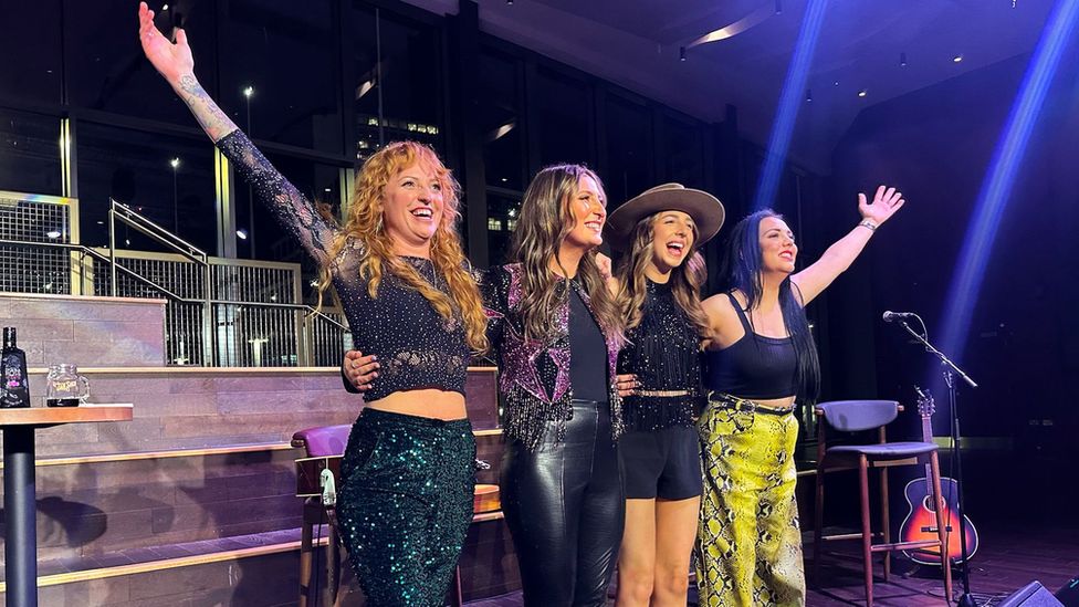 L-R Kezia Gill, Jess Thristan, Demi Mariner, and Jade Helliwell. The four women are pictured on stage, hugging each other and smiling while Kezia and Jade hold their arms out. They all have long hair and are dressed in sparkly outfits - Demi is also wearing a cowboy hat. Behind them are four chair and a guitar and spotlights lighting the stage which is otherwise dark.
