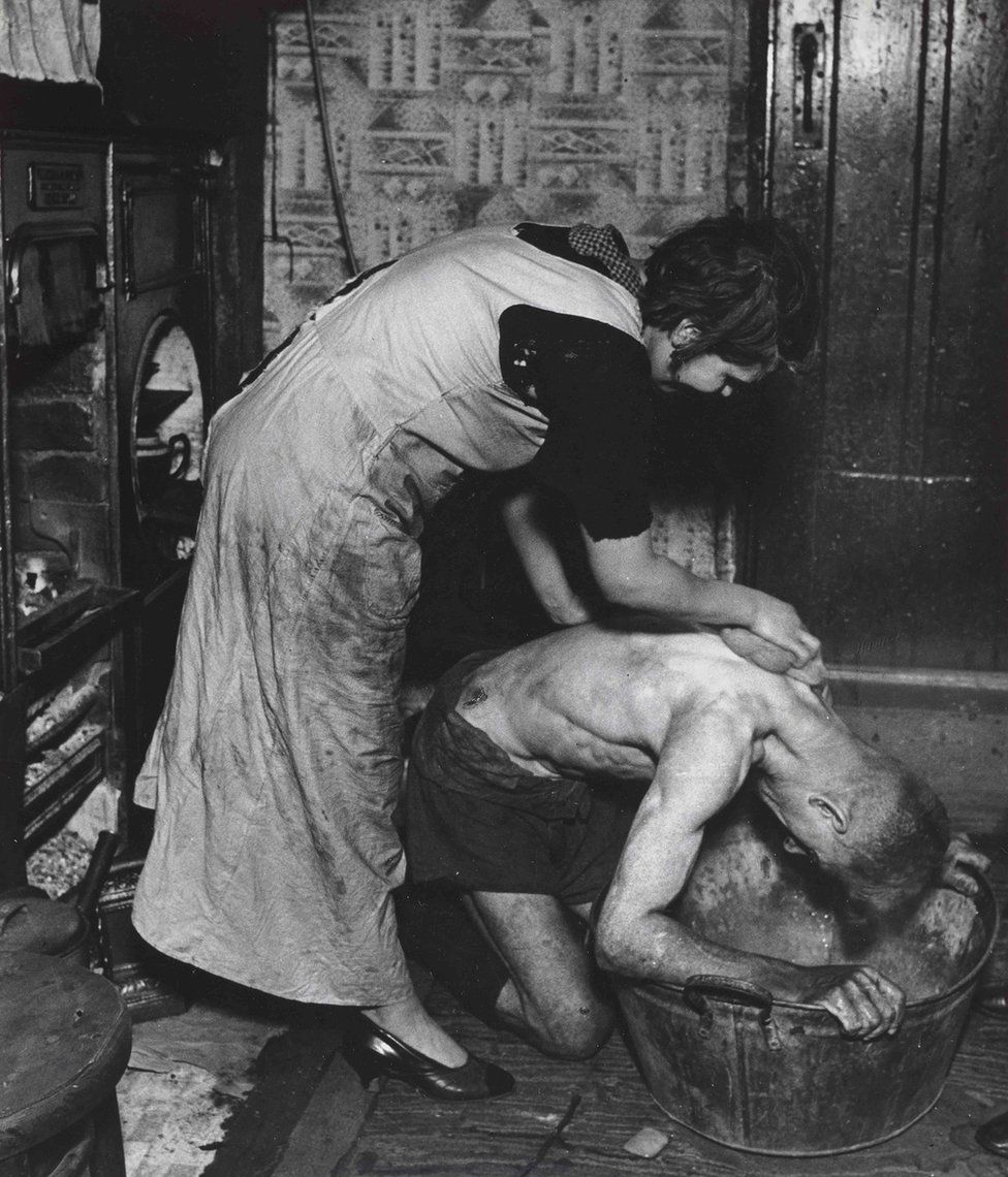 A photograph of a woman helping a man bathe in a tin bath
